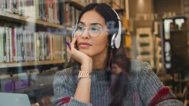 Woman in library wearing headphones, looking thoughtful.