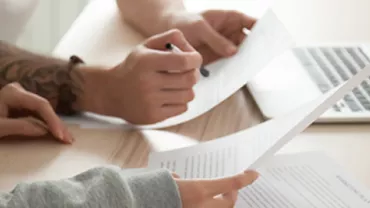 Two people reviewing documents with a laptop on a desk.