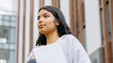 Young woman with long hair, holding a laptop, standing confidently outside an office building.