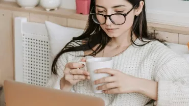 Woman with glasses sipping coffee, using a laptop on a cozy couch.