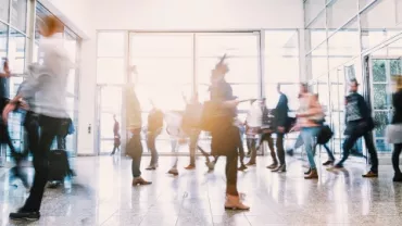 Blurred motion of people walking through a busy, bright lobby.