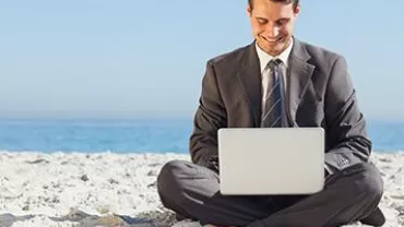 Man in suit working on laptop while sitting on a beach.