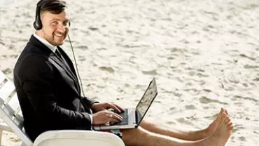 Man in a suit with headphones working on a laptop on the beach.