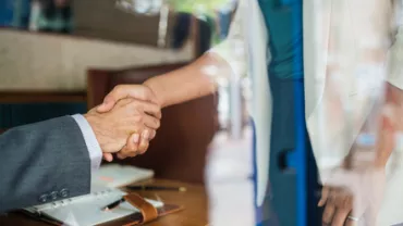 Two people shaking hands over a desk in an office setting.