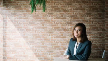 Woman in a suit sitting at a table with a brick wall background.