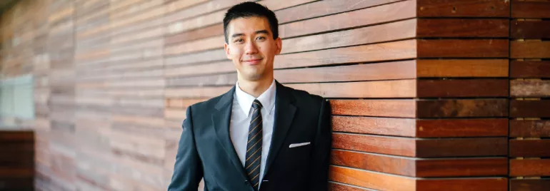 Smiling man in a suit leaning against a wooden wall.