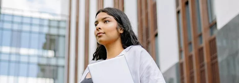 Young woman with long hair, holding a laptop, standing confidently outside an office building.