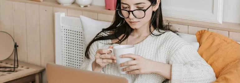 Woman with glasses sipping coffee, using a laptop on a cozy couch.